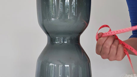 Person holding a measuring tape next to a dark gray vase on a light surface with a neutral background