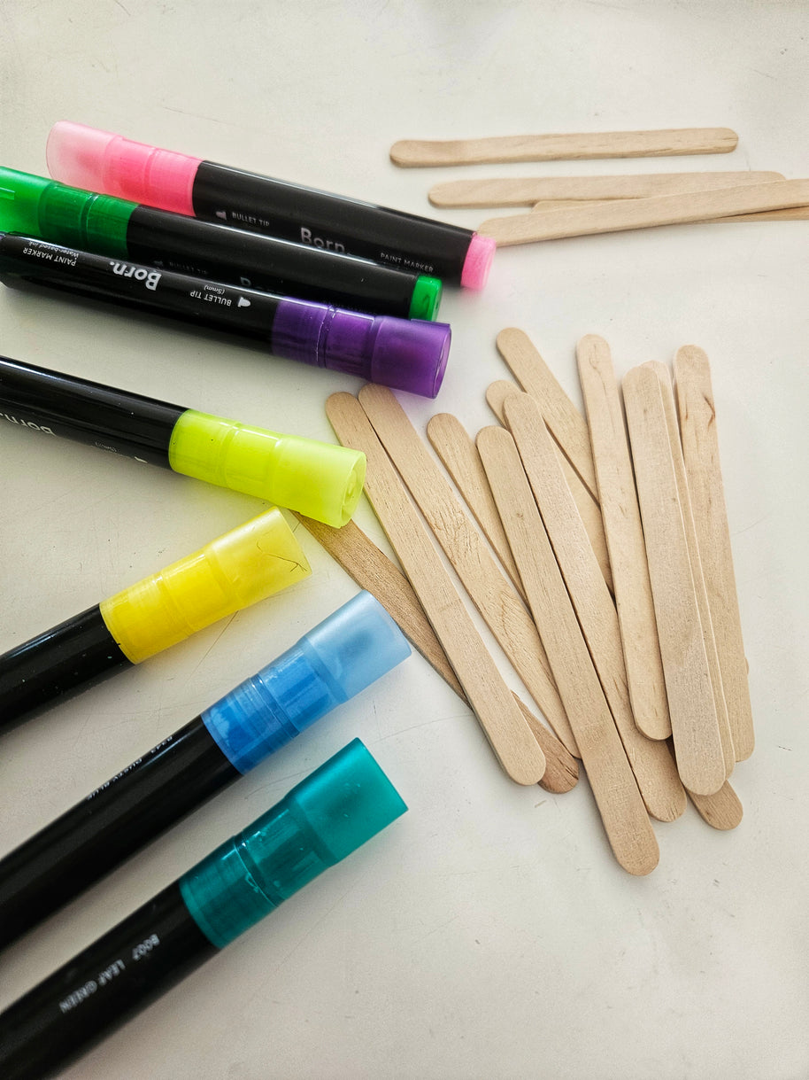 Colourful paint markers and wooden popsicle sticks on a white surface