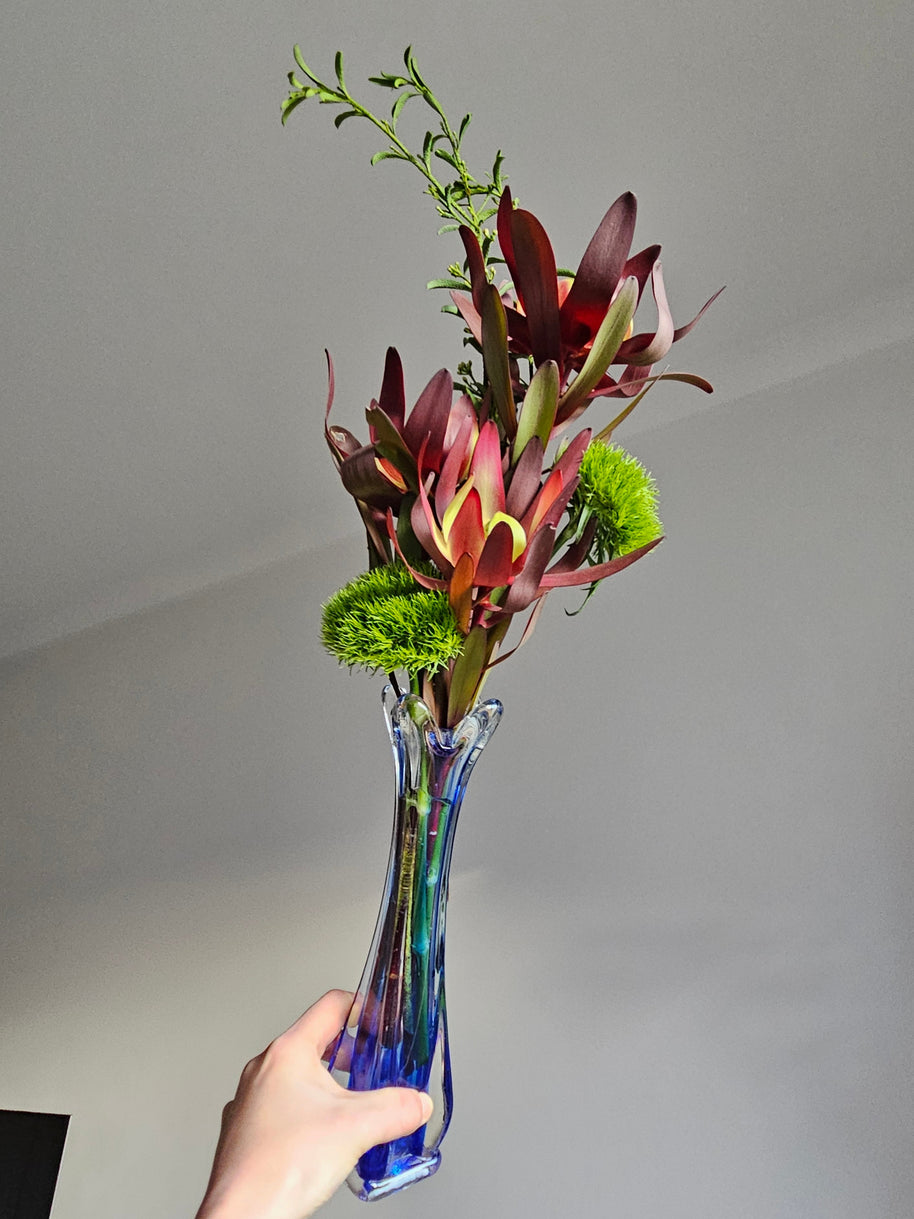 Hand holding a blue glass vase with a bouquet of flowers against a plain background