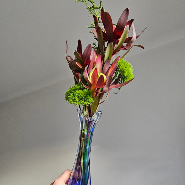 Hand holding a blue glass vase with a bouquet of flowers against a plain background