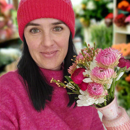 Woman wearing a pink knit hat and sweater, holding a bouquet of flowers in a floral shop.