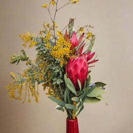 Bouquet of flowers with red venus protea and yellow wattle in a red vase against a beige background