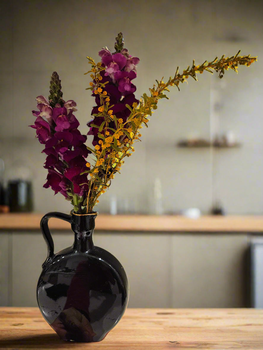 Black vase with purple and yellow flowers on a wooden table in a kitchen setting