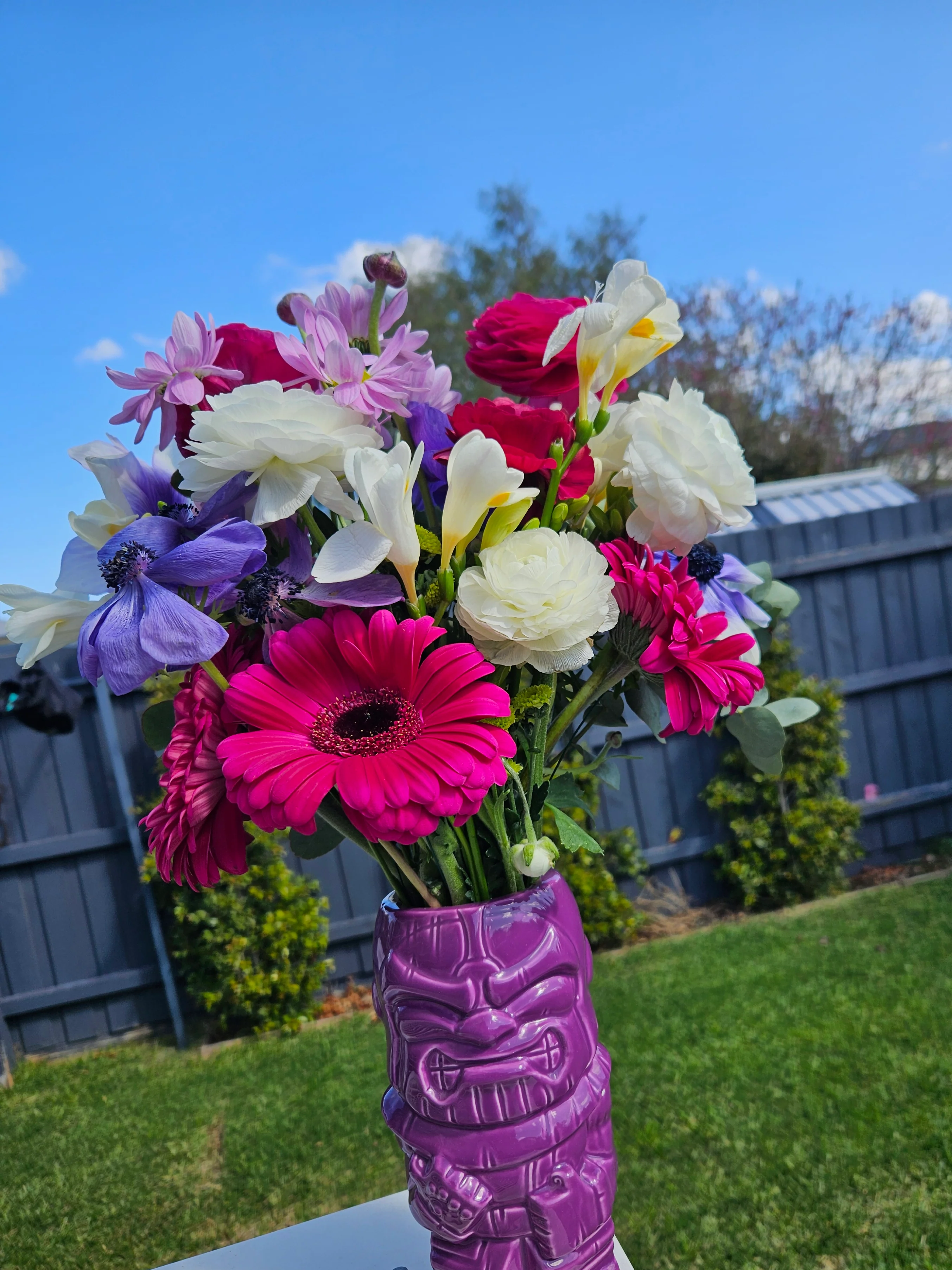 Bouquet of colourful flowers in a purple tiki mug vase outdoors.