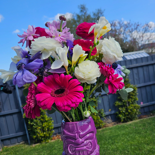 Bouquet of colourful flowers in a purple tiki mug vase outdoors.