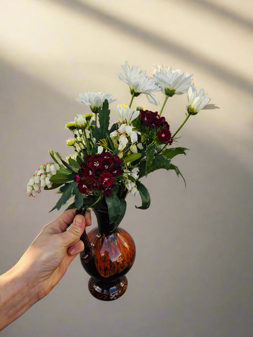 Hand holding a small brown jug vase with flowers against a plain background