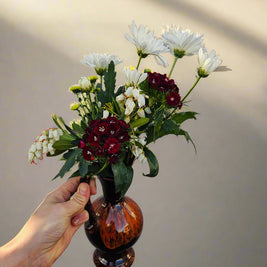 Hand holding a small brown jug vase with flowers against a plain background