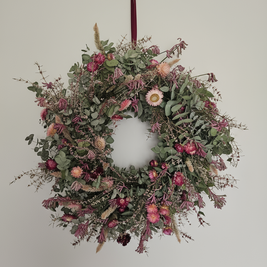 Decorative pink and green Christmas wreath with dried flowers and foliage hanging on a plain wall.