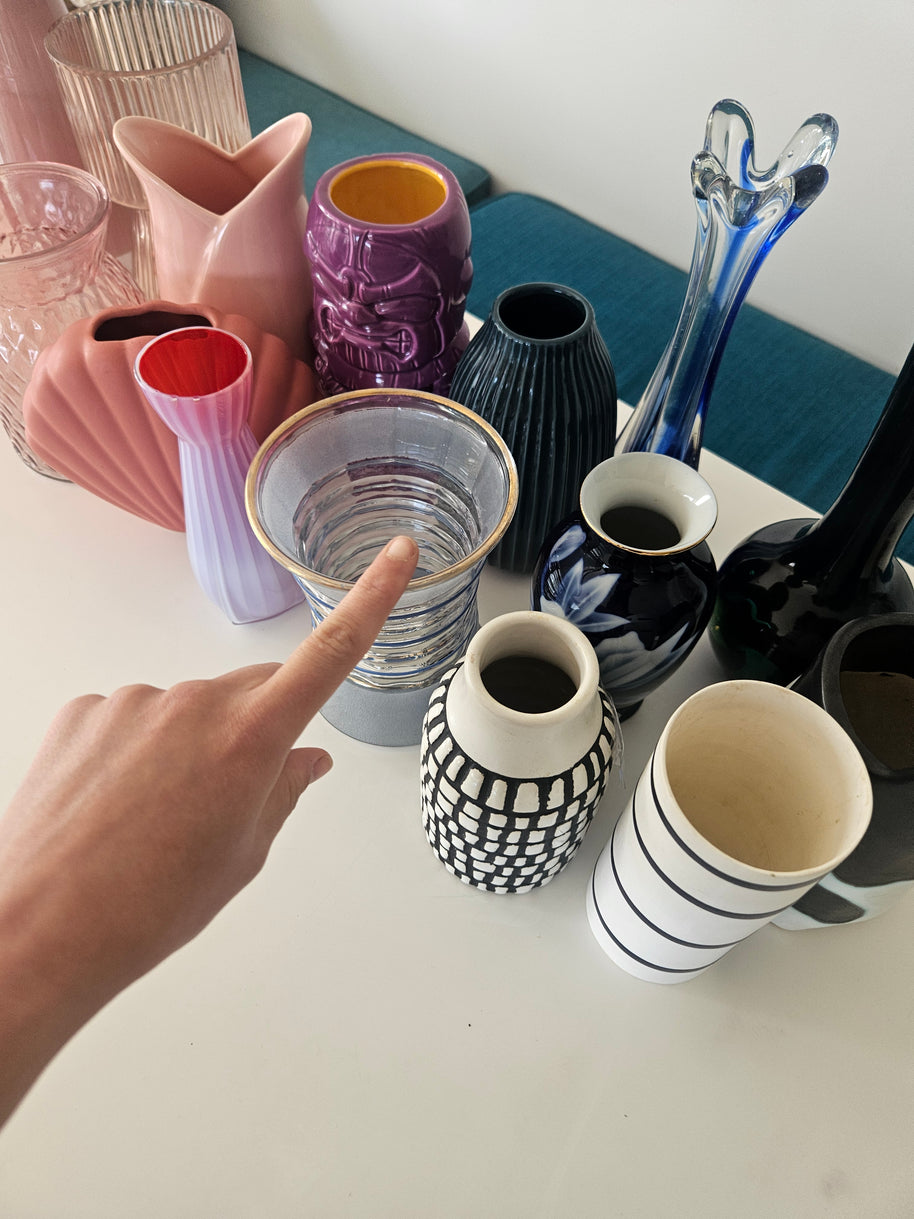 Hand pointing at a clear glass vase among various colourful vases on a table.