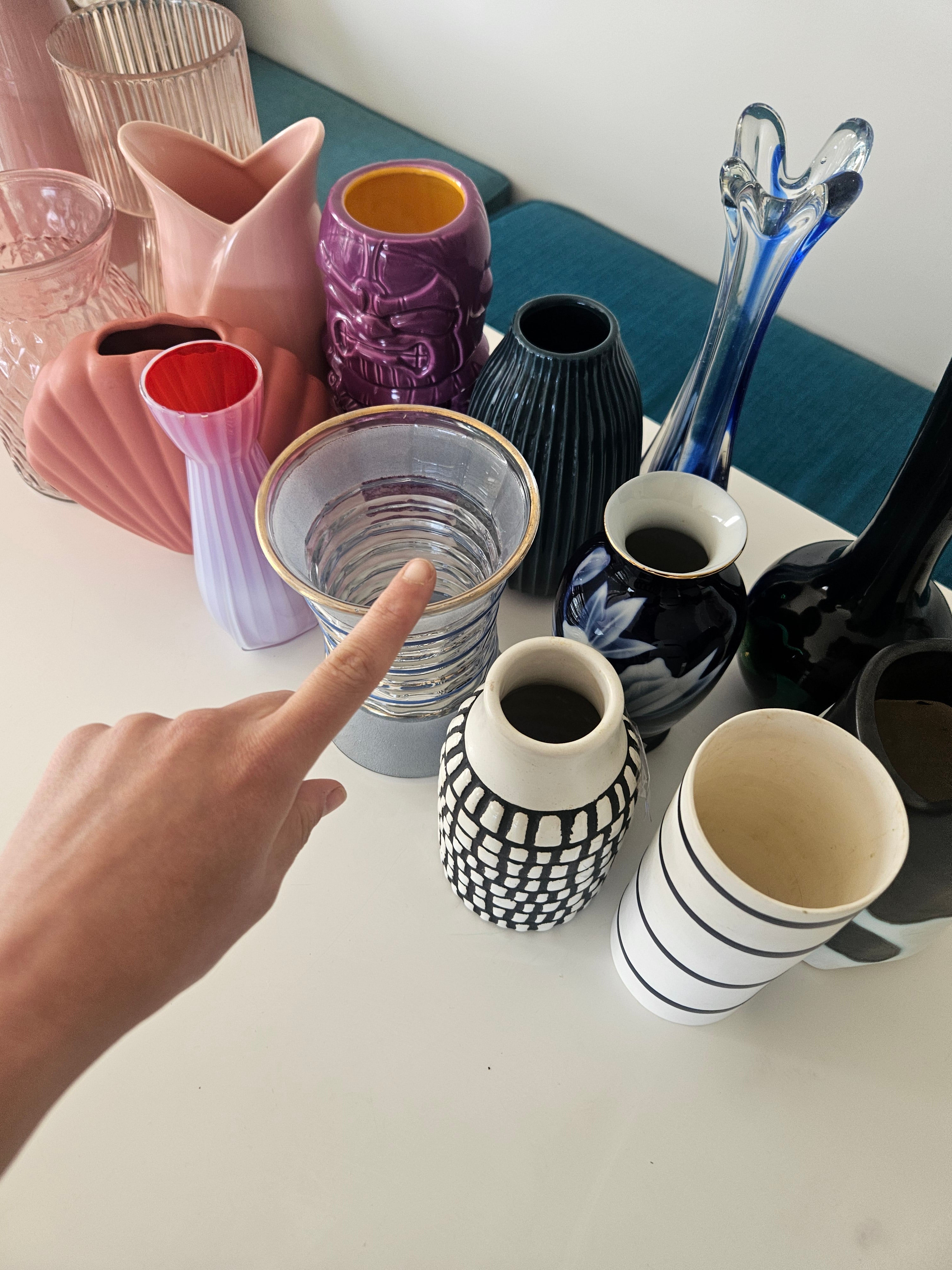 Hand pointing at a clear glass vase among various colourful vases on a table.
