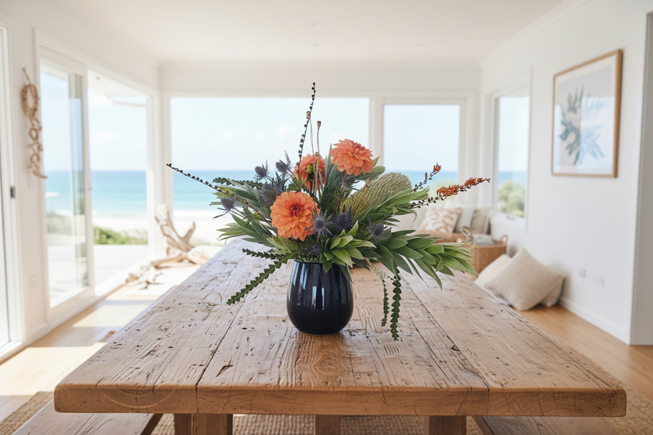 Floral arrangement with orange dahlias, banksia and green leaves in a black vase against a white background