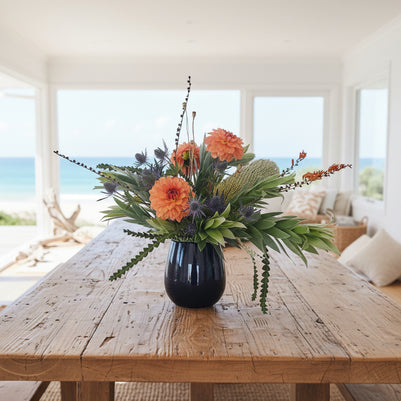 Floral arrangement with orange dahlias, banksia and green leaves in a black vase against a white background