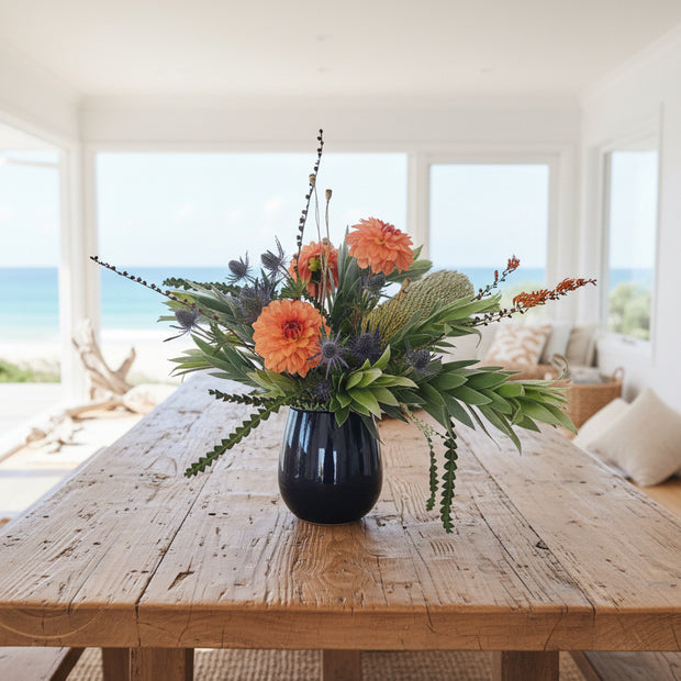 Floral arrangement with orange dahlias, banksia and green leaves in a black vase against a white background