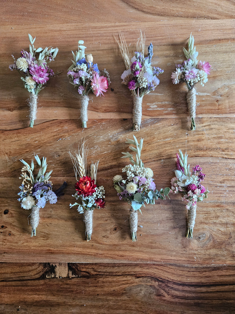 Eight small dried floral boutonniere wristlets arranged in two rows on a rustic wooden surface. The bouquets contain mixed pastel and earthy tones including pink, purple, blue, red, beige, and green, with some wheat stems and greenery.