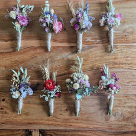 Eight small dried floral boutonniere wristlets arranged in two rows on a rustic wooden surface. The bouquets contain mixed pastel and earthy tones including pink, purple, blue, red, beige, and green, with some wheat stems and greenery.