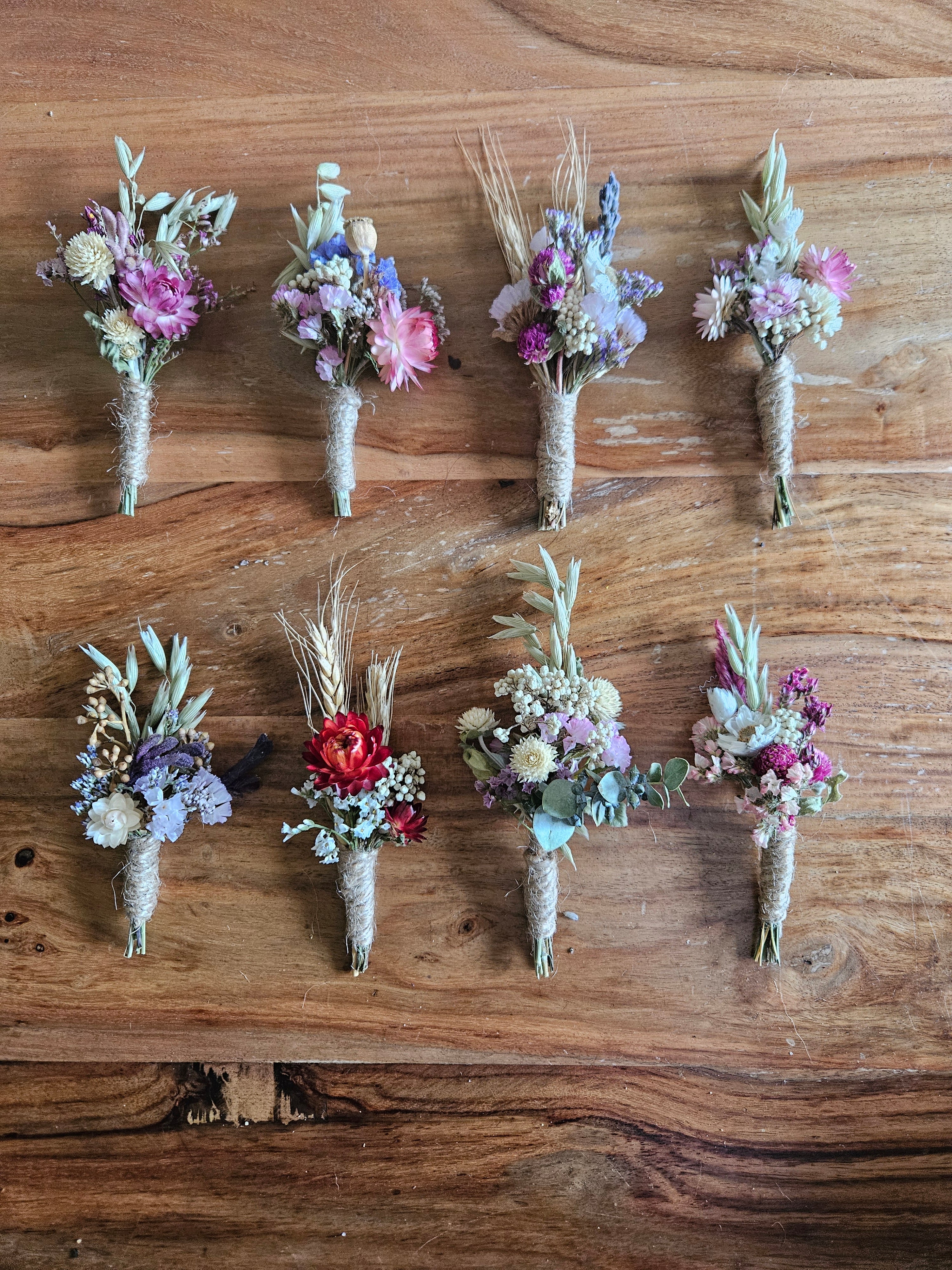 Eight small dried floral boutonniere wristlets arranged in two rows on a rustic wooden surface. The bouquets contain mixed pastel and earthy tones including pink, purple, blue, red, beige, and green, with some wheat stems and greenery.