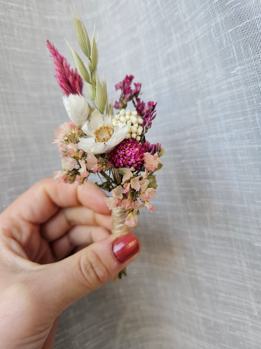 A hand with red nail polish holds a small dried floral boutonniere featuring pink, white, and purple flowers against a textured light white fabric background.