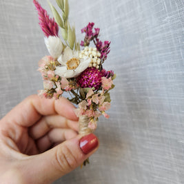 A hand with red nail polish holds a small dried floral boutonniere featuring pink, white, and purple flowers against a textured light white fabric background.