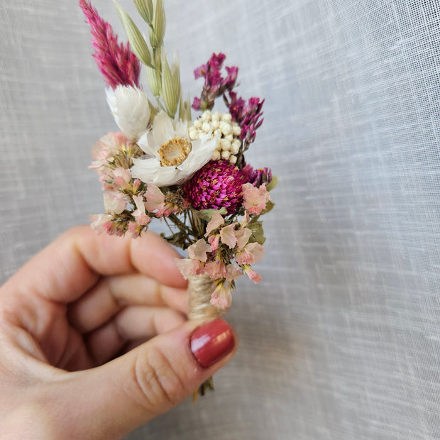 A hand with red nail polish holds a small dried floral boutonniere featuring pink, white, and purple flowers against a textured light white fabric background.