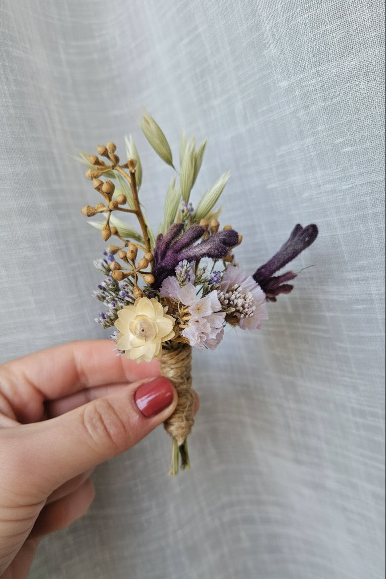 A hand with pink nail polish holds a small dried flower boutonniere featuring cream, purple, and beige botanicals against a textured light gray fabric background.
