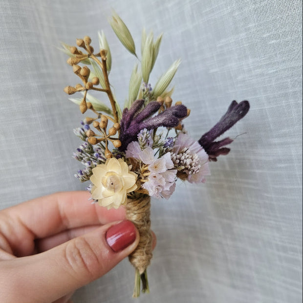 A hand with pink nail polish holds a small dried flower boutonniere featuring cream, purple, and beige botanicals against a textured light gray fabric background.