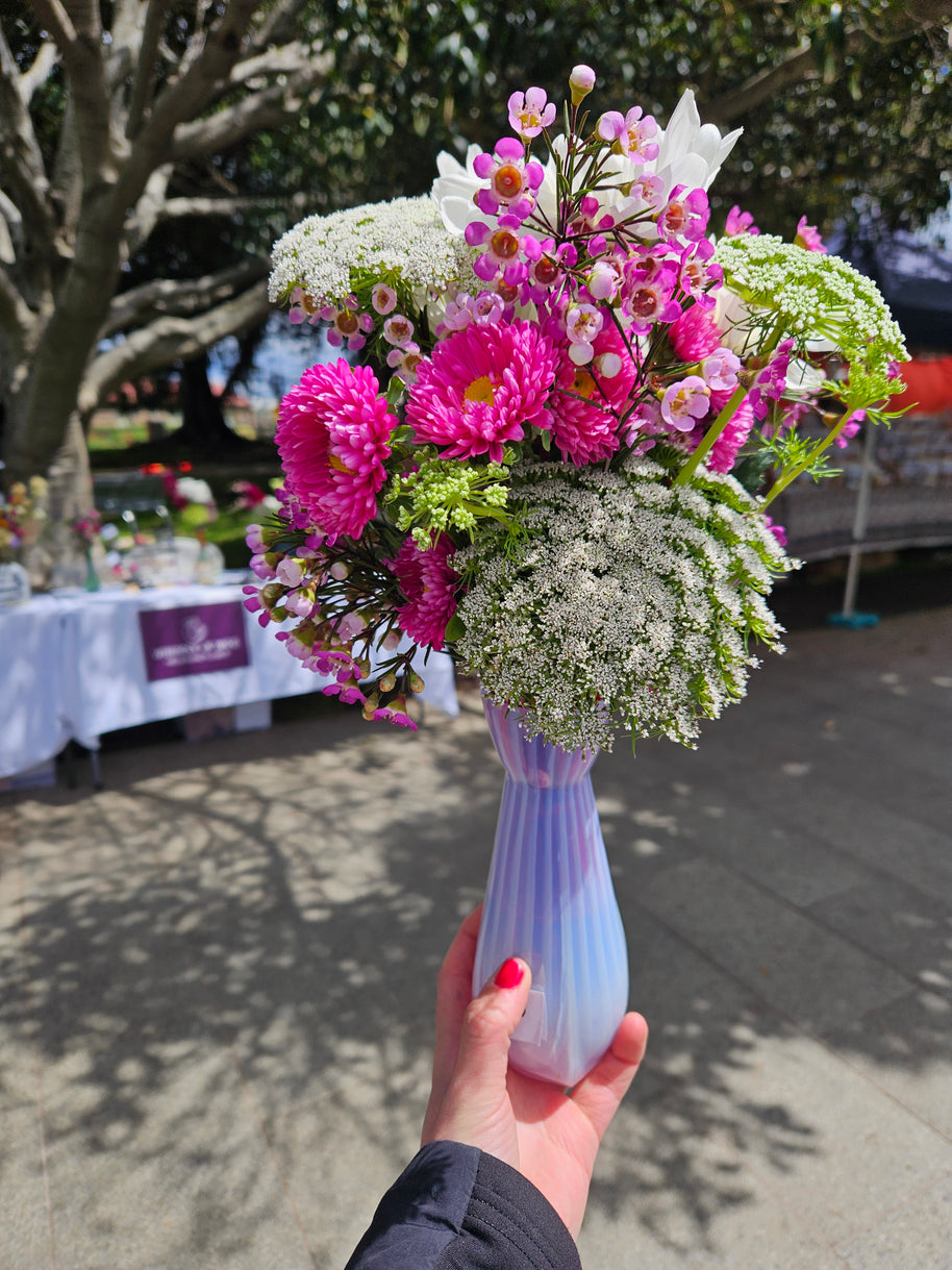 Person holding a pink and white flowers in a pink glass vase outdoors at a market