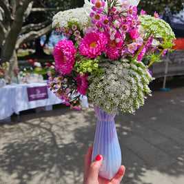 Person holding a pink and white flowers in a pink glass vase outdoors at a market