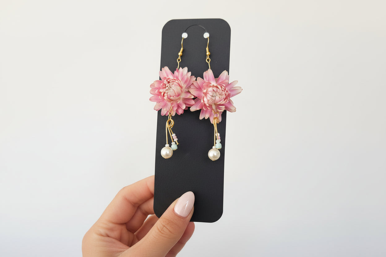 Pink floral earrings with gold and pearl details on a black card held by a hand.