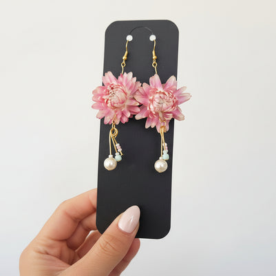 Pink floral earrings with gold and pearl details on a black card held by a hand.