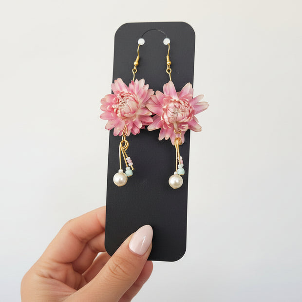 Pink floral earrings with gold and pearl details on a black card held by a hand.